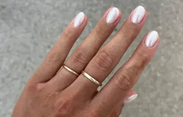Dłoń z delikatnymi, złotymi pierścionkami i eleganckimi paznokciami perłowymi, lśniącymi subtelnym blaskiem.