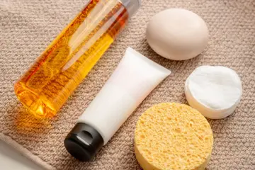 Płyn do kąpieli z **sodium lauroyl sarcosinate**, krem, mydło i waciki kosmetyczne na ręczniku.